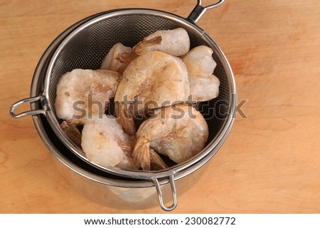 Frozen shrimp in strainer dripping into stainless steel mixing bowl on wooden cutting board.