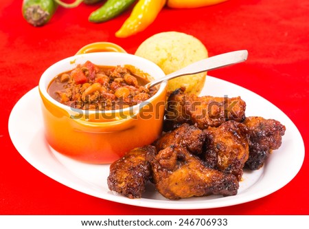 Serving of spicy wings in Honey BBQ Sauce on plate with bowl of homemade chili and Jalapeno cornbread..  Bright red tablecloth with copy space.