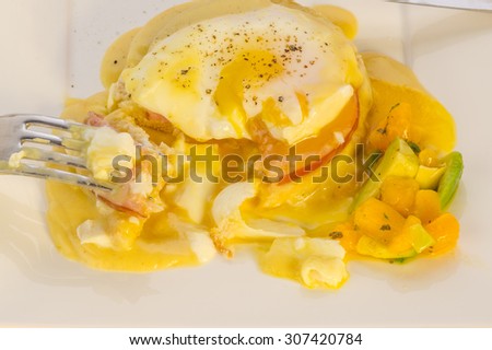 Overhead view of bite taken from Perfectly poached egg over bacon on English Muffin.  Topped with creamy Hollandaise Sauce and served with spicy  fruit salad.