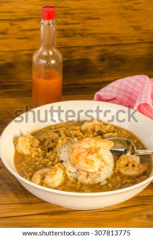 Shrimp Gumbo in large white bowl with bottle of Louisiana Hot Sauce on old wooden picnic table with pink gingham napkin.  Vertical format.