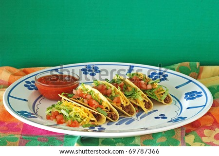 Five tacos on large platter with bowl of salsa, all in colorful setting.