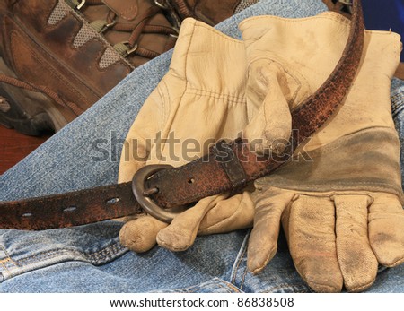 Heavy work boots with old leather belt and dirty work gloves on faded blue denim background signify the uniform of hard working men.