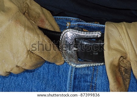 Man\'s hand in leather gloves with leather belt and silver buckle on denim jeans to conceptually represent an ranch worker or ranch hand in cowboy talk.