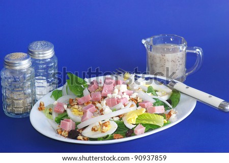 Sliced boiled eggs and chopped ham over bed of lettuce with onions and feta cheese with Fat Free Poppy Seed Dressing against blue background.