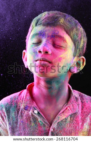 A boy plays Holi with colored powder exploding around his face in a dark background.
