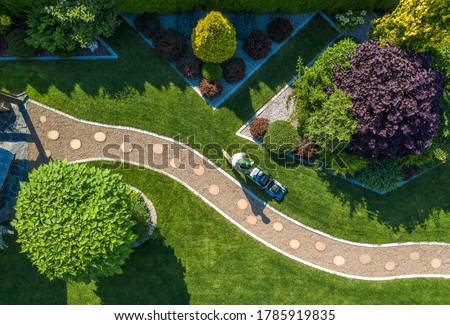 Caucasian Gardener with Grass Mower Trimming Beautiful Backyard Garden Lawn. Aerial View. Gardening and Landscaping Industry.