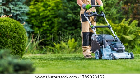 Professional Caucasian Gardener in His 40s Trimming Grass Lawn Using Modern Electric Cordless Mower. Landscaping Industry Theme.