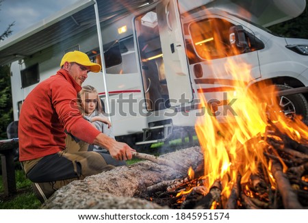 Recreational Vehicle RV Camper Camping and Family Time. Caucasian Father and His Daughter Hanging Next to Campfire on Their RV Park Pitch. Class C Motorhome in Background.