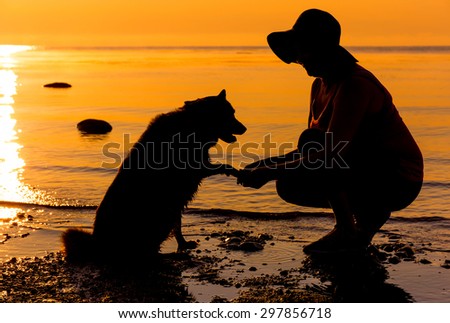 Lady in hat and dog shaking hands on sea shore at sunrise time