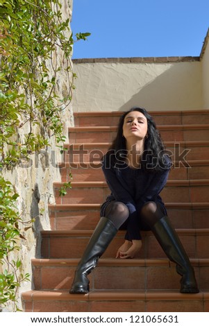 Stylish beautiful girl sits on the stairs near rough stone wall of the old house under bright sun. Tuscan, Italy