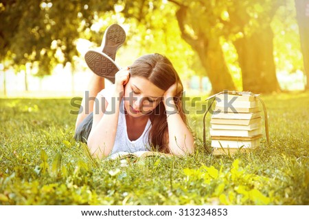Young beautiful college student girl lying down on the green grass and reading a book at campus at warm day. Education. Back to school conceptual image.