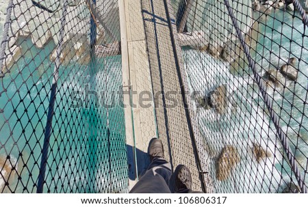 Booted feet of man walking on narrow wooden swing bridge crossing over clear fresh white water rushing through a rocky gorge
