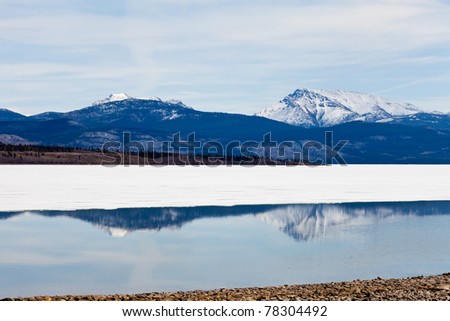 First open water at thawing Lake Laberge, Yukon Territory, Canada: reflection of snow-covered mountains on calm open water surface of still largely ice-covered lake.