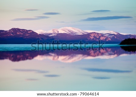 The evening before ice-break at Lake Laberge, Yukon Territory, Canada: reflection of snow-covered mountains on calm open water surface of still largely ice-covered lake.