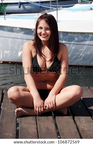Beautiful young woman in black bikini in yacht harbor. Happy girl sitting on the bridge with crossed legs and smiling in front of yacht boat.