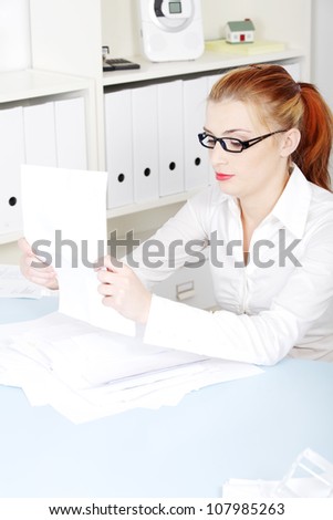 Young businesswoman sitting at desk and organizing files, documents and papers. Blond girl in black glasses and white shirt is doing paper work