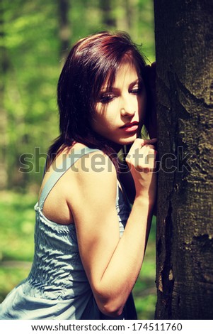 Young woman holding face next to tree trying to hear voice of nature. Girl standing with ear next to wood. Pretty girl with eyes closed and summer dress.