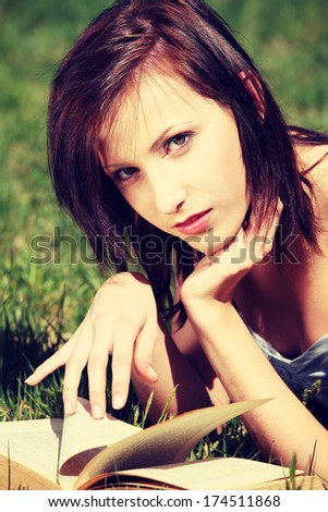 Young woman during warm sunny day, wearing dress, lying on grass relaxing and with open book. Young girl with open holding book with one hand holding her chin and by another flipping pages.