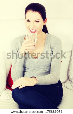 Front view portrait of a young beautiful smiling woman sitting on a sofa, holding a glass of water next to her slips.