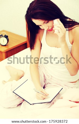 Picture of a full lenght woman, sitting on a bed, arranging a meeting through the phone - alarm clock in the background.