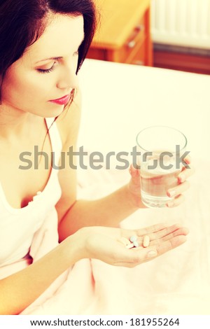 Site view of a beautiful young woman in bed, holding pills and a glass of water.