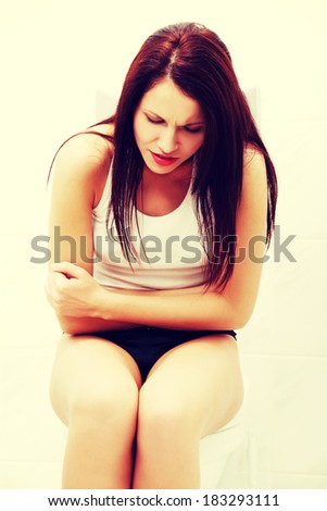 Front view of a suffering woman because of the belly ache, sitting in bathroom embracing her stomach.