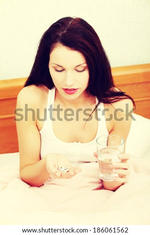 Front view prtrait of a beautiful young woman in bed, holding pills and a glass of water.
