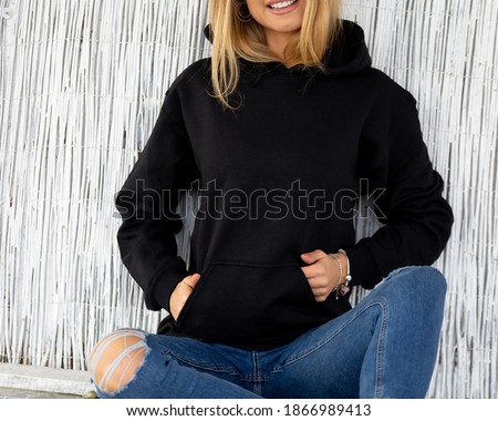 Unrecognizable woman sits in front of camera, she wears black hoodie. There is a place on hoodie for inscription or design for fashion mockup. Casual outfit for lady.  Sweatshirt template.