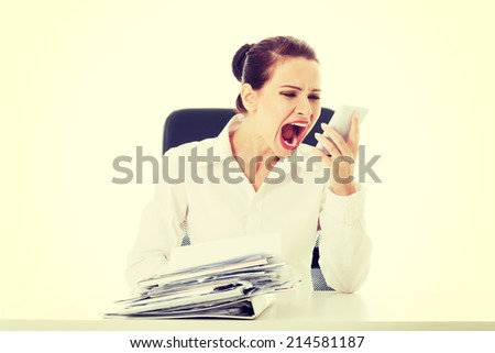 Beautiful business woman sitting, screaming to a phone and has a stack of papers on desk. Isolated on white.