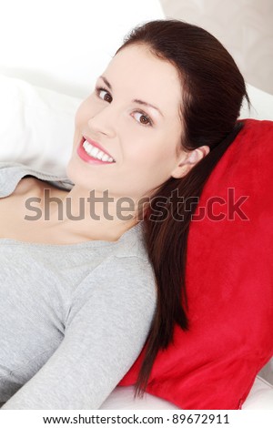 Site view portrait of a young beautiful smiling woman lying on a couch, resting her head on a red pillow, looking opposite to the camera.