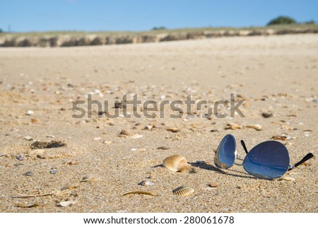 A pair of tinted sunglasses on beach sand.Shallow focus on foreground. There are some seashells scattered around the beach. Background has out of focus surf and island. Sun glare on sun-glass frame.