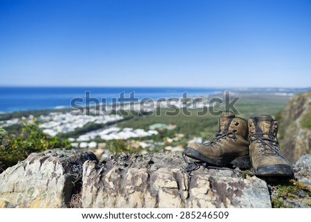 A pair of old hiking boots on top of a mountain. Shallow focus on foreground with boots and rocks. Background has out of focus settlement and ocean view with blue sky.