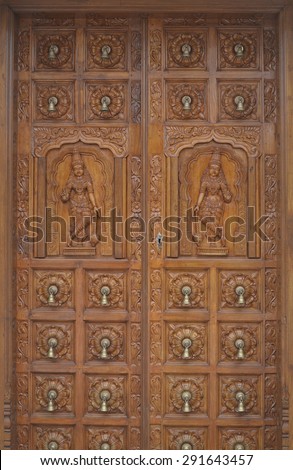 Traditional Hindu wood carvings on large double timber door. The doors are studded with small brass bells arranged in a grid pattern Symmetrical female figures on each leaf of the doors on the middle.