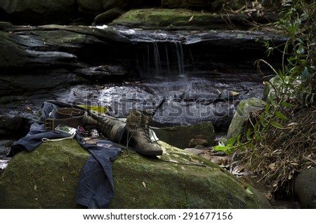 A hiker\'s boots and personal belongings on a moss covered rock by a forest creek. Hiking gear consists of shirt, dark cargo pants, worn out hiking boots, headlamp, compass, leather belt and knife.