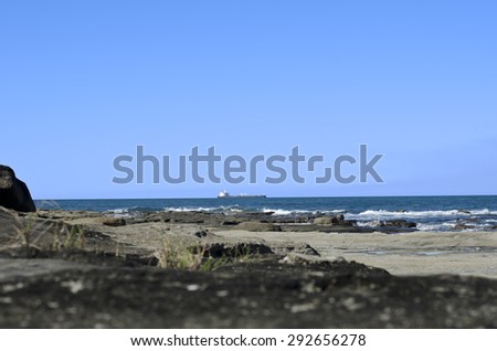 Large container cargo ship on ocean. Foreground has rocky shore and breaking waves. Background has blue sky. Image taken near Caloundra, Queensland Australia.