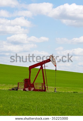 An oil well with the pump jack in action, against a grassy, green hill & cloudy blue sky.  Located in the province of Alberta, Canada.