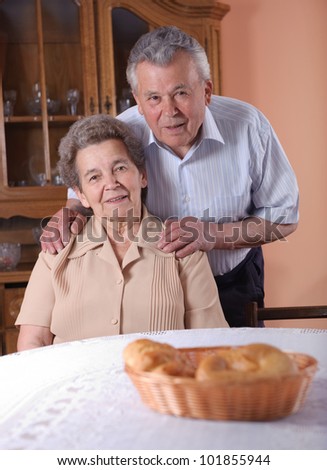 Happy senior couple at the table indoors. Hands on shoulders.