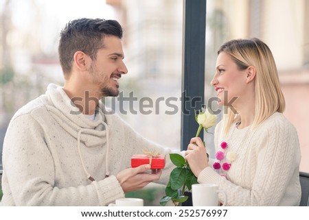 Romantic young couple sitting in a restaurant. Young man holding gift box, woman holding white rose.