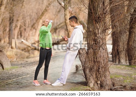 Two young people resting after physical activities in nature. Young woman drinking water and young man using mobile phone.