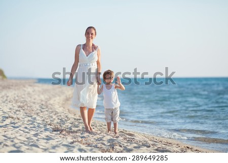 Happy young mother walking along the beach with her little son waving to camera