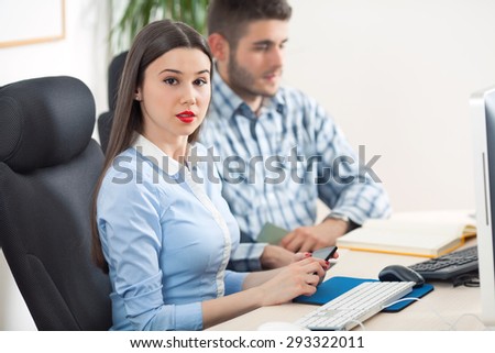 Young businesswoman is sitting in an office in front of the computer holding mobile phone and looking at camera. Young businessman in the background is sitting next to her.