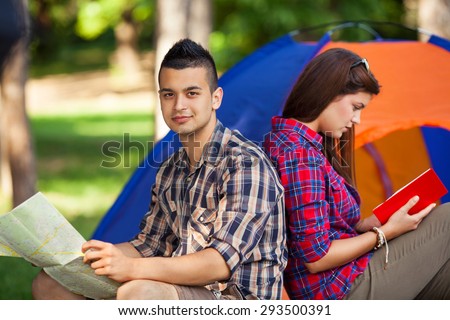 Young man sitting with his girlfriend in front of the tent in a forest, looking at maps and planning their camping adventure