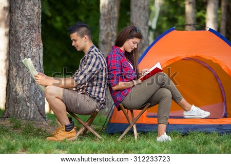 Teenage couple sitting back to back in front of the tent, young man looking at maps while girl is reading a book