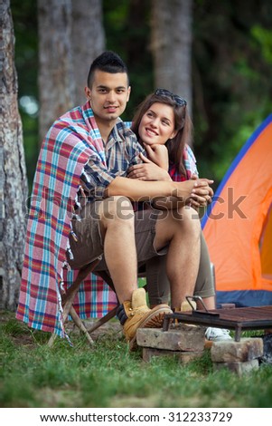 Young camping couple wrapped in blanket warming up by the fire in front of the tent