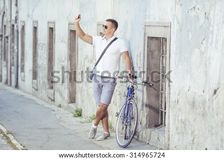 Young man standing on the old town street with his bicycle beside him and taking a photo with his smart phone