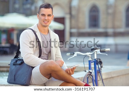 Smiling young businessman sitting in the street next to his bicycle and using digital tablet