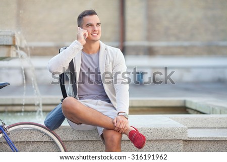 Portrait of smiling young man talking on mobile phone while sitting near the fountain in the city