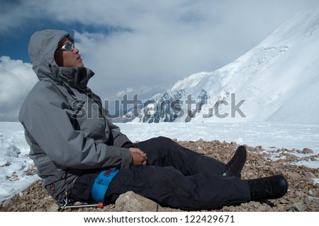 Climber looking at the Lenin peak while having rest on his way up. Ice bore is attached to his belt for self-rescue means. Branches and slopes of Trans-Alay Range can be seen on the background.