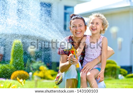 Portrait of little gardener girl with mother watering flowers on the lawn near cottage.