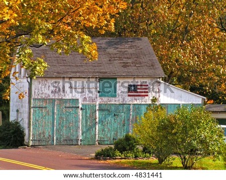Small old white barn with aqua doors and peeling paint.  A folk style American Flag adorns the side of the barn.  Beautiful autumn setting.